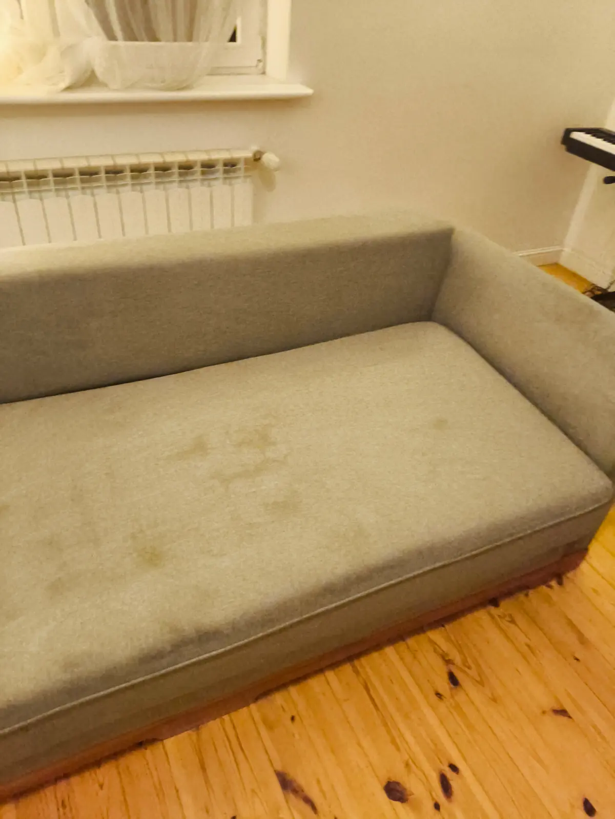 Szara sofa z zaokrąglonymi podłokietnikami przed praniem — ciemne plamy na siedzisku. Pranie kanapy Grudziądz, SofaGo