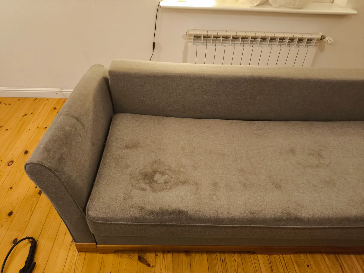Szara sofa z zaokrąglonymi podłokietnikami przed praniem — ciemne plamy na siedzisku. Pranie kanapy Poznań, SofaGo