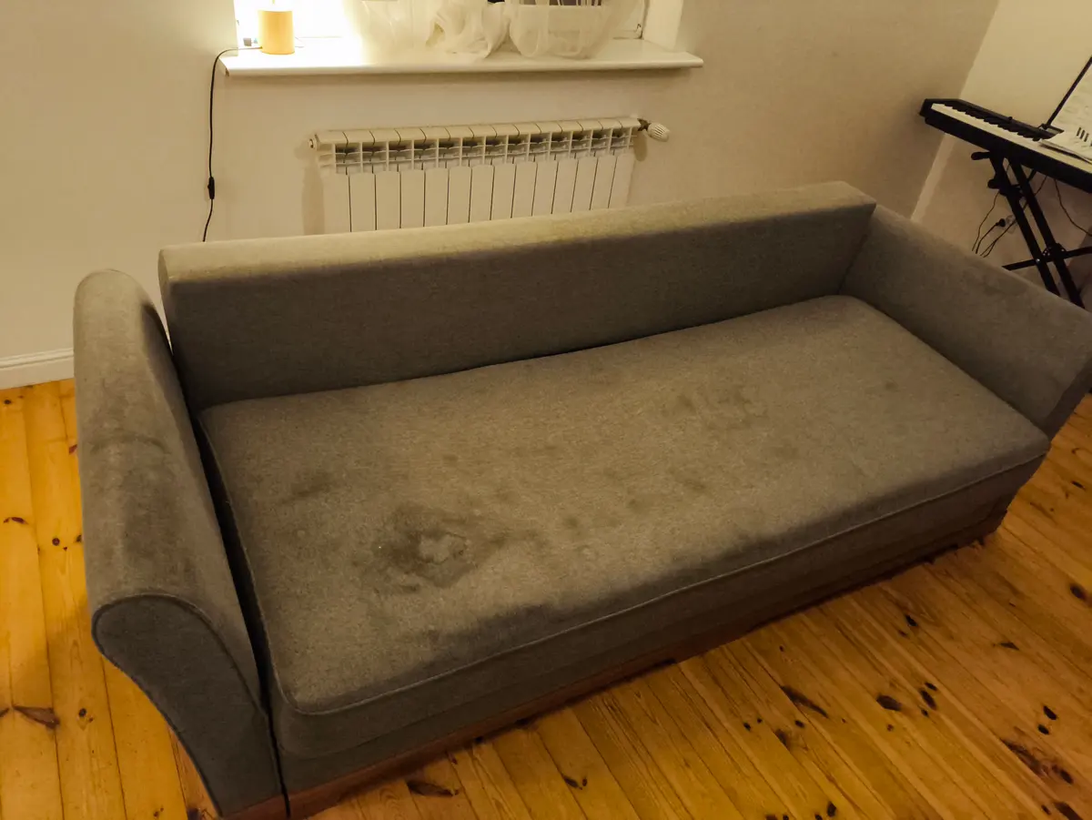 Szara sofa z zaokrąglonymi podłokietnikami przed praniem — ciemne plamy na siedzisku. Pranie kanapy Szczecin, SofaGo