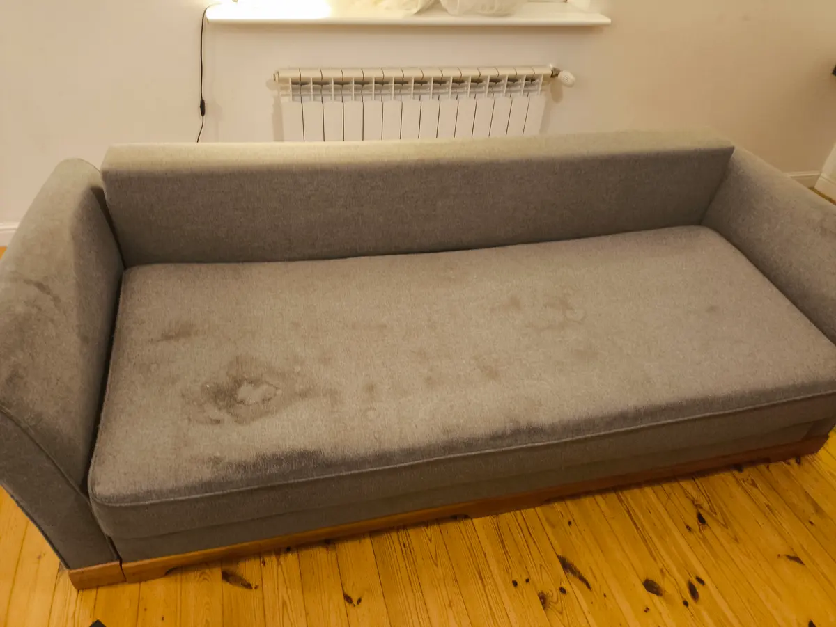 Szara sofa z zaokrąglonymi podłokietnikami przed praniem — ciemne plamy na siedzisku. Pranie kanapy Warszawa, SofaGo